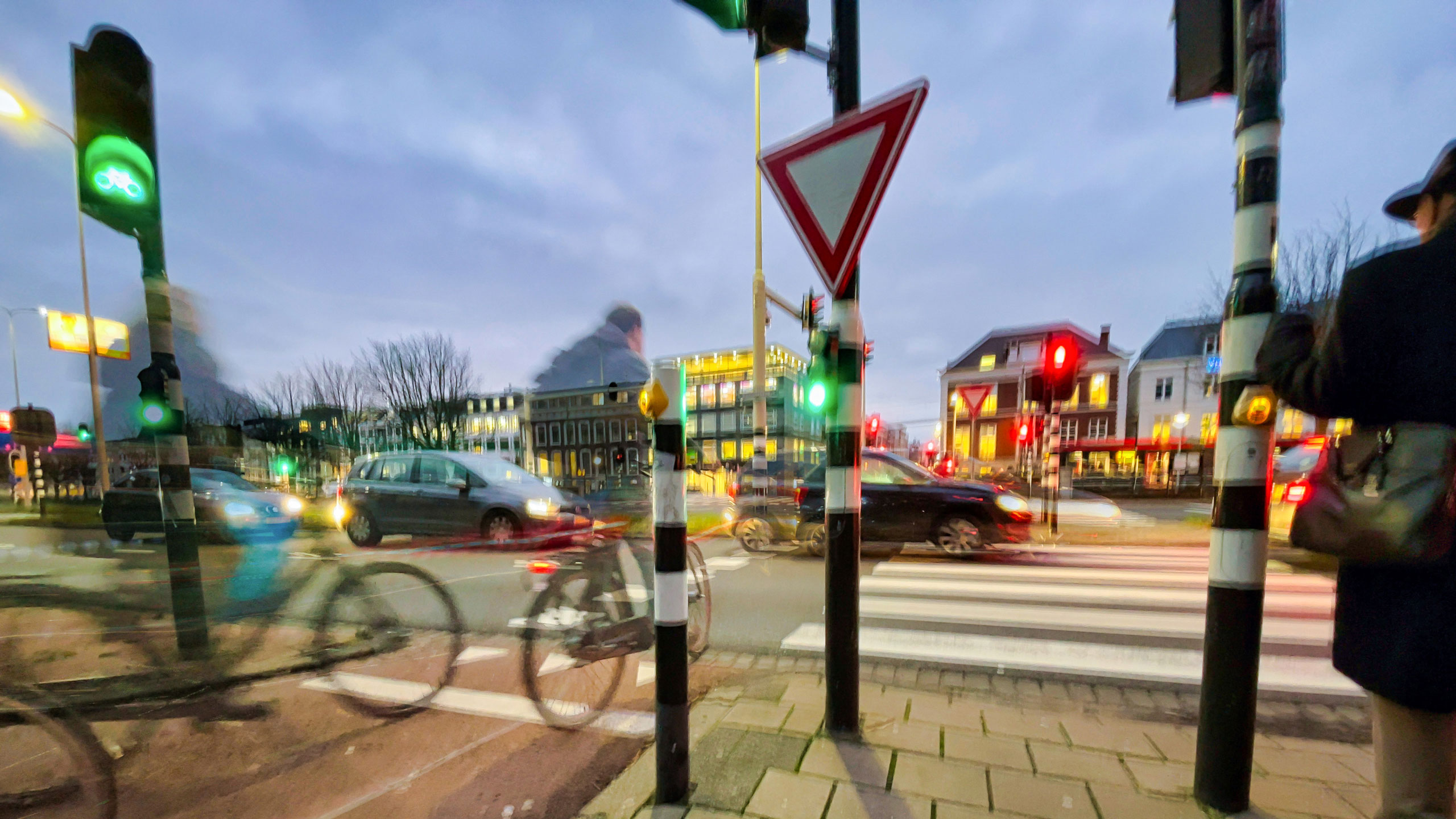 In de schemering: Kruispunt gezien vanaf een fietspad met daarop een aantal fietsers. Voor de fietsers is er groen licht, maar op het kruispunt staan wel auto’s.