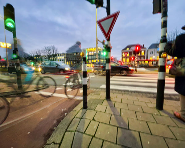 In de schemering: Kruispunt gezien vanaf een fietspad met daarop een aantal fietsers. Voor de fietsers is er groen licht, maar op het kruispunt staan wel auto’s.